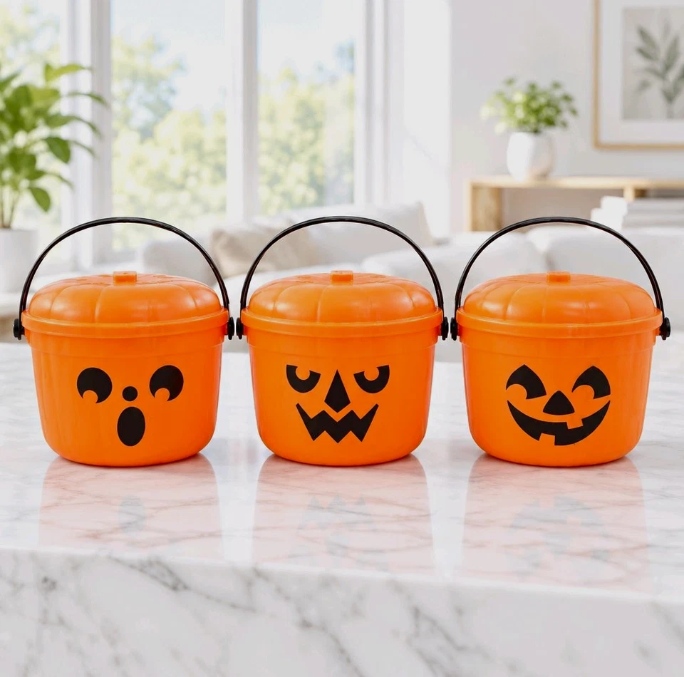 Three orange plastic buckets with pumpkin-shaped lids, black handles and different black faces stand on a pale stone counter in a bright room.