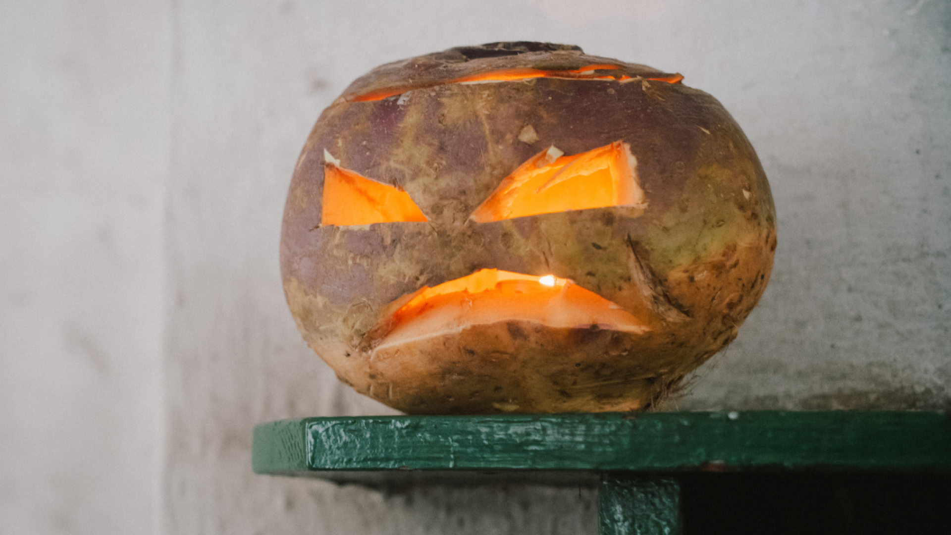 Squat brown and purple root vegetable carved with angular eyes and a downturned mouth, glowing orange from within on a green shelf against a pale wall.