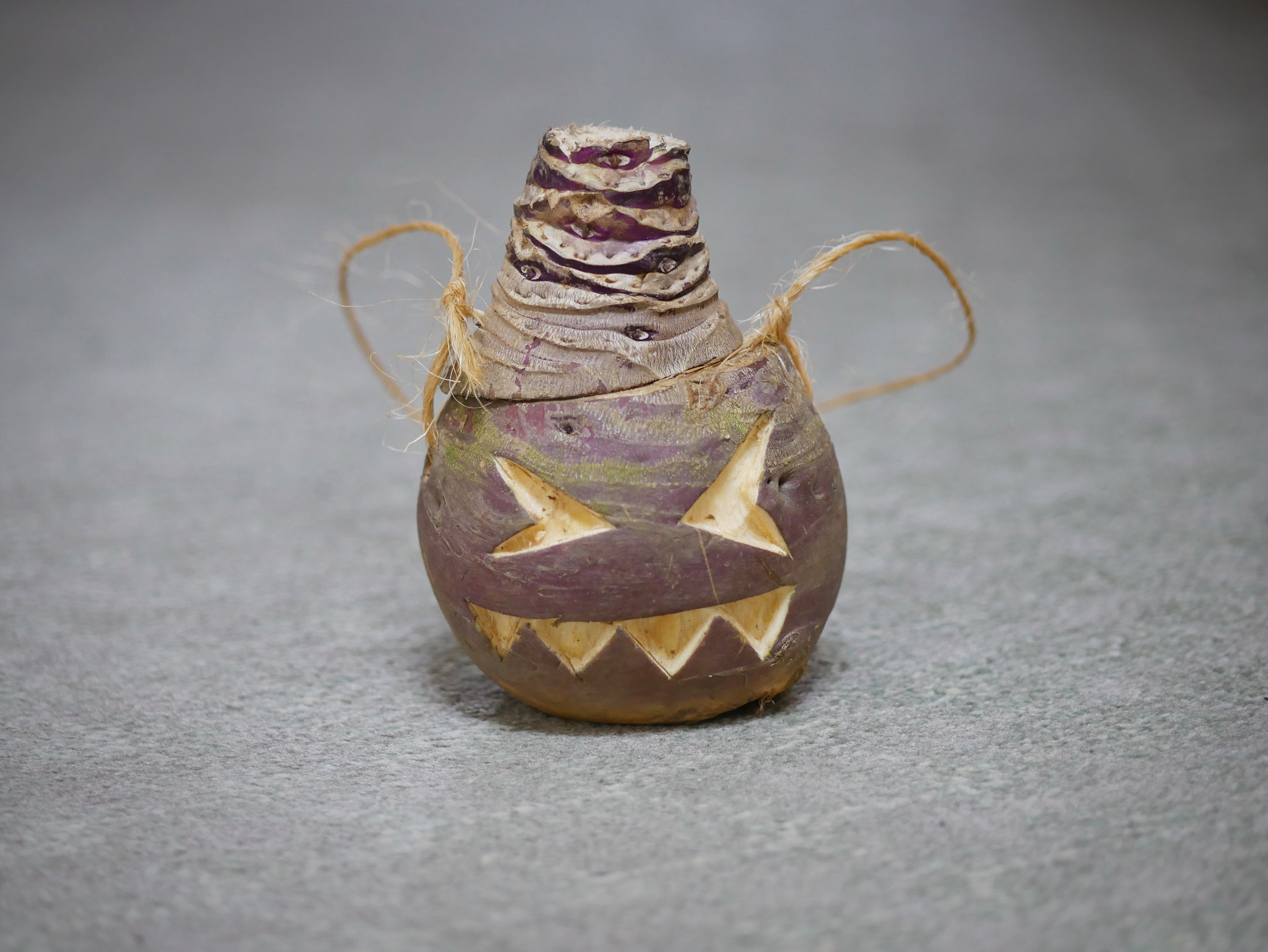 Purple and cream root vegetable carved as a lantern with star-shaped eyes, a wide jagged-toothed mouth, stacked cut sections on top and a looped twine handle, standing against a grey background.