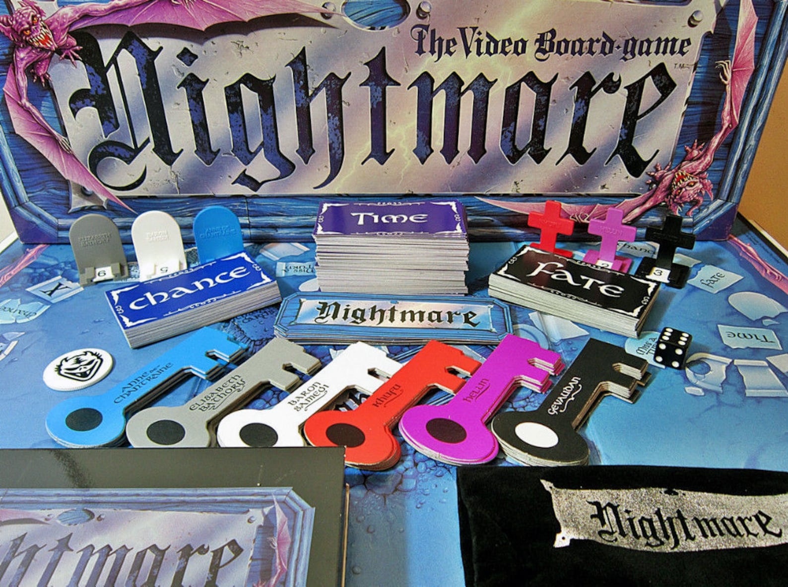 Close view of a blue Nightmare game board with the branded box lid behind stacks of Time, Chance, Fate and Nightmare cards, coloured key-shaped counters, tombstone pawns, cross-shaped pieces, a black die and a cloth pouch.