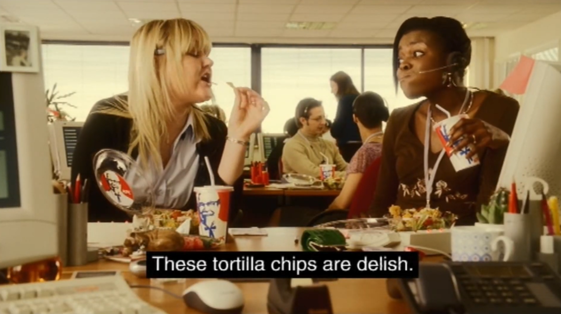 Two women wearing telephone headsets sit at desks in a busy office, eating from KFC containers. Computers, telephones and other workers fill the background, and a subtitle reads, “These tortilla chips are delish.”