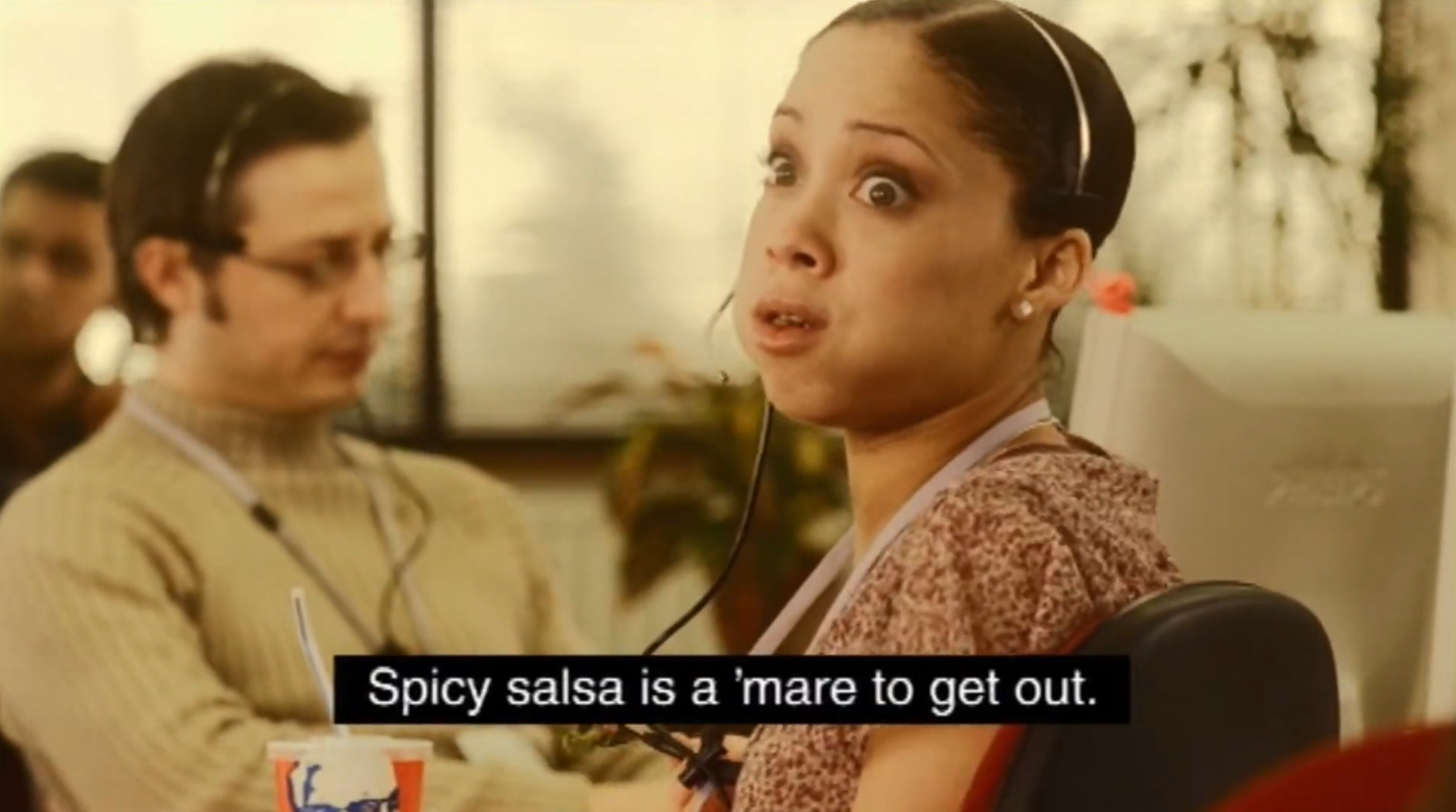 Call-centre worker wearing a headset holds a KFC cup beside a colleague. The subtitle reads: “Spicy salsa is a ’mare to get out.”
