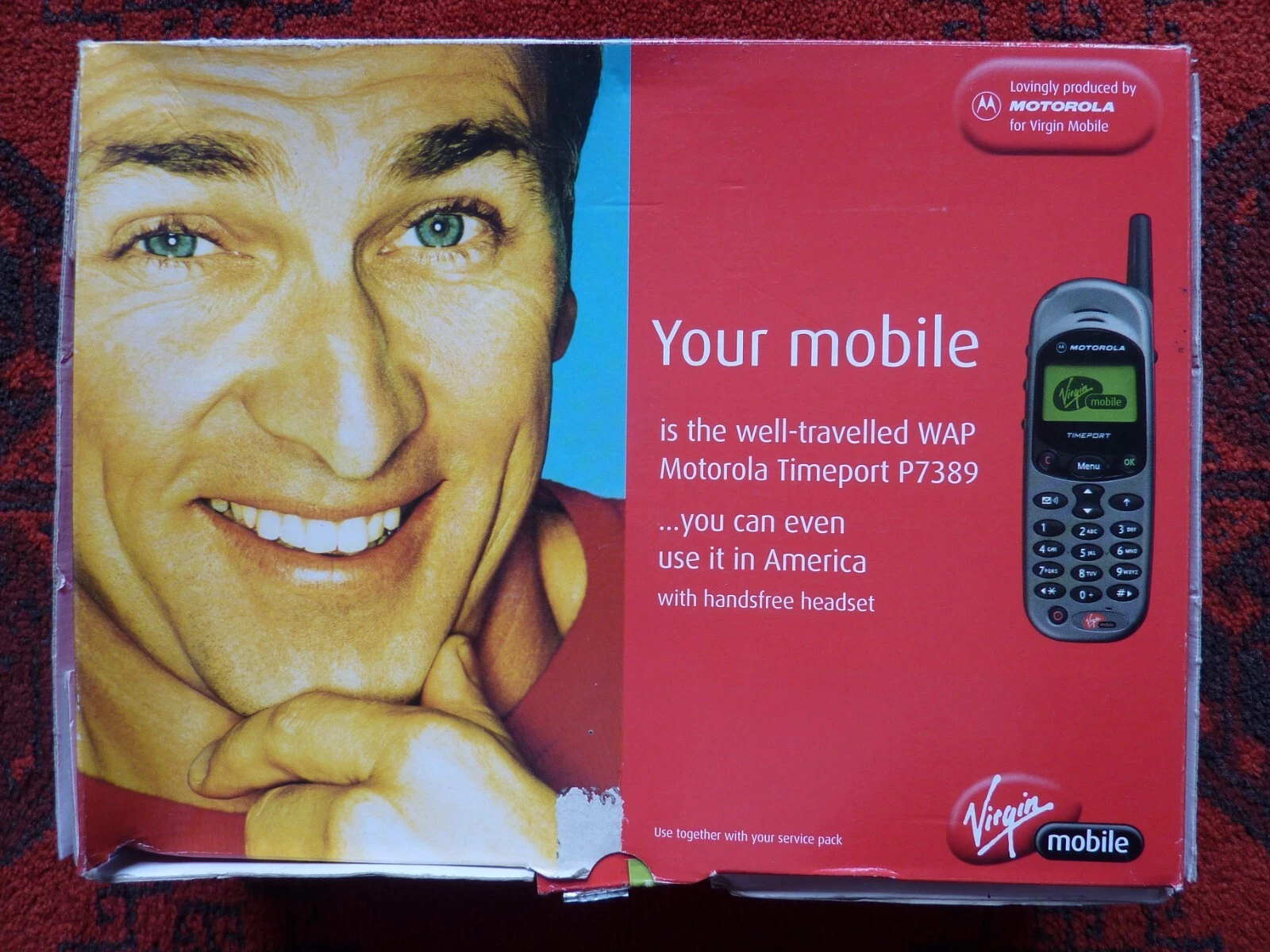 Worn red Virgin Mobile printed spread on patterned carpet, with a smiling man on the left and a Motorola phone on the right. Text identifies it as the WAP Motorola Timeport P7389, and the phone screen displays the Virgin Mobile logo.