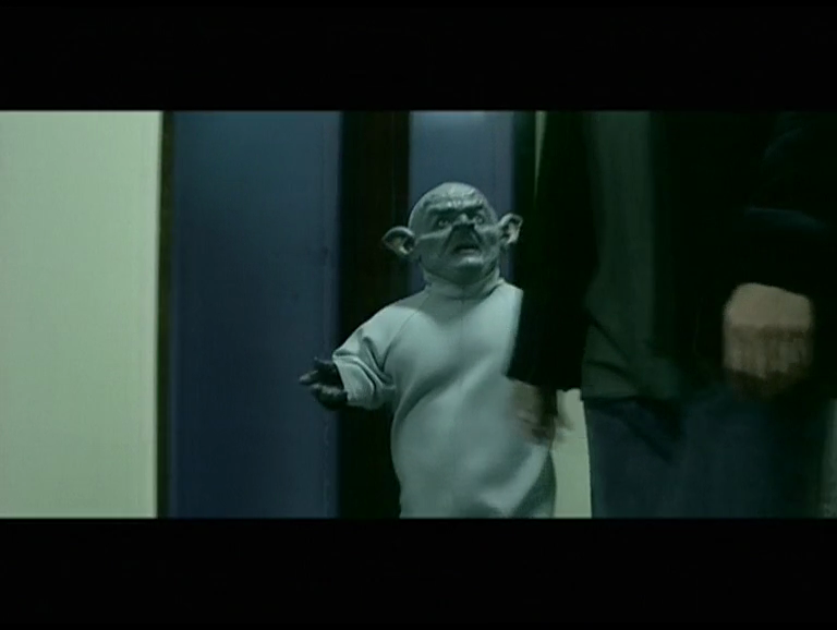 Grey, pointed-eared Gremlin standing beside a man in a doorway, in a letterboxed television still.