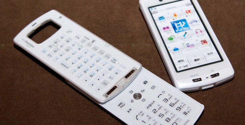 White modular mobile phone on a brown surface, with the touchscreen display detached beside a keyboard section opened to show QWERTY and numeric keypads.