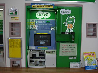 Front view of a green-and-white ticket machine built into a station wall, with a central screen, payment and ticket slots, two Kaeru logos, and a large cartoon frog on the right panel. Smaller wall units and a leaflet rack flank the machine.