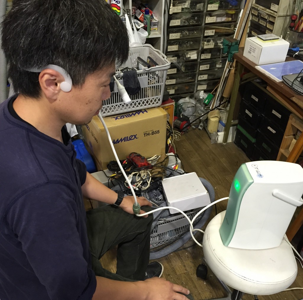 Man seated in a workshop wearing a white ear-mounted headset connected by tubing to a pale-green and white oxygen air charger with an illuminated indicator.