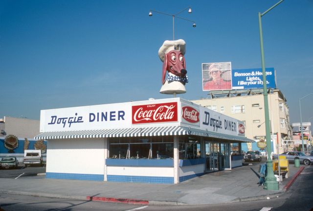 Colour photograph of a white-and-blue Doggie Diner restaurant with striped awnings, Coca-Cola signs and a giant dachshund head wearing a chef's hat on the roof.