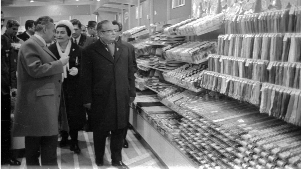 Black-and-white photograph of a formally dressed group walking beside long shop counters densely stocked with hanging packets, folded goods and rows of small packaged items.