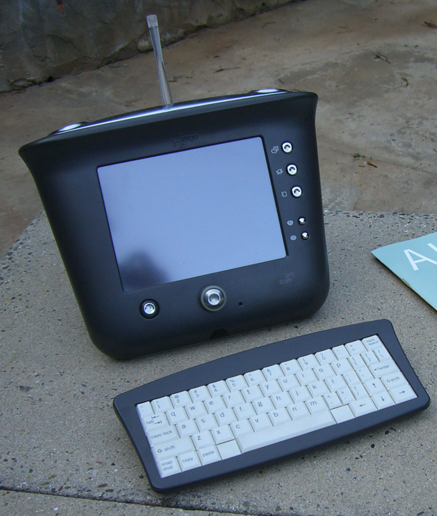 Dark 3Com Ergo Audrey viewed from the front and left on a concrete surface, with a blank reflective screen, upright stylus, circular dial and compact keyboard.