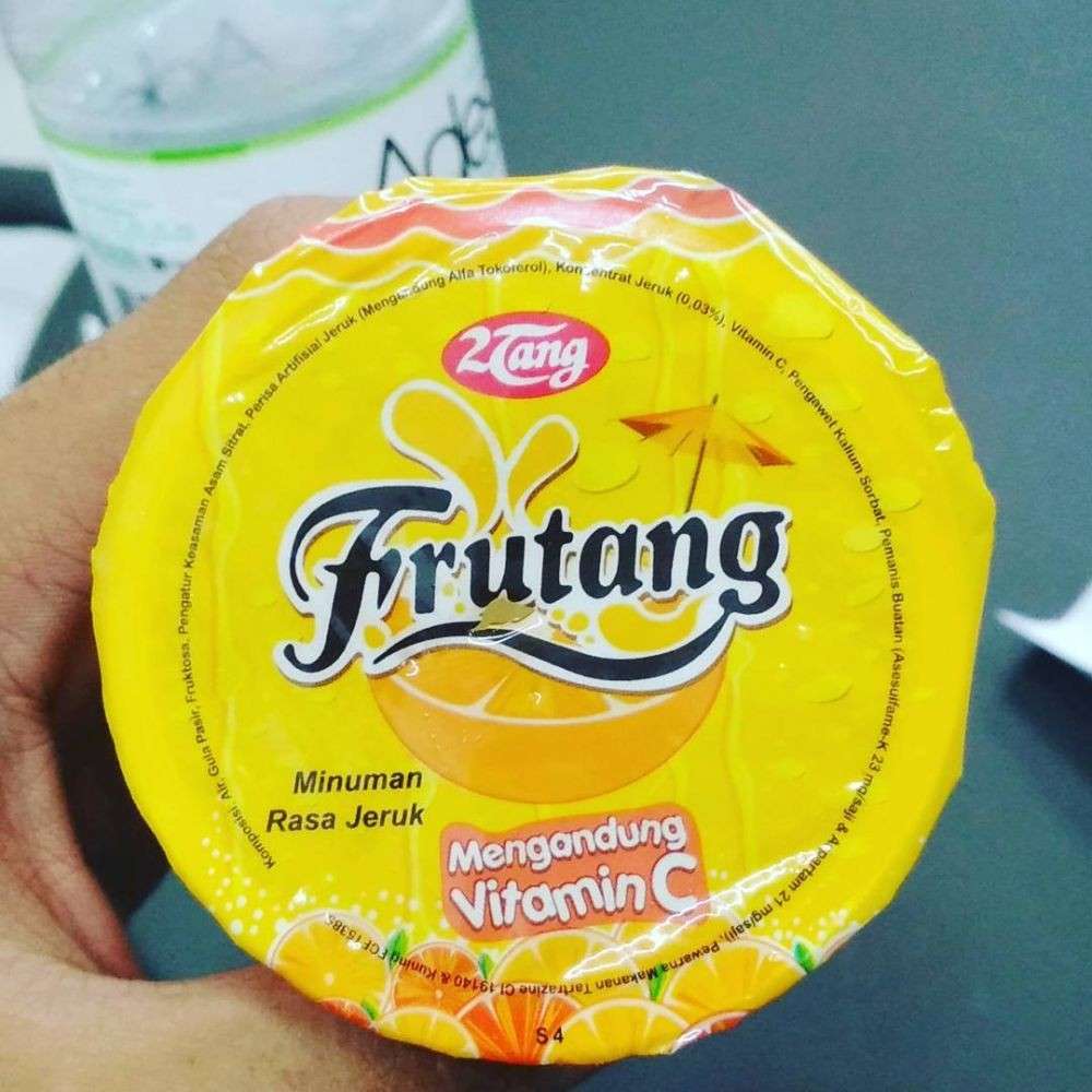 Close-up of a yellow Frutang orange drink foil lid held in a hand, with the 2Tang logo, orange illustrations and vitamin C branding.