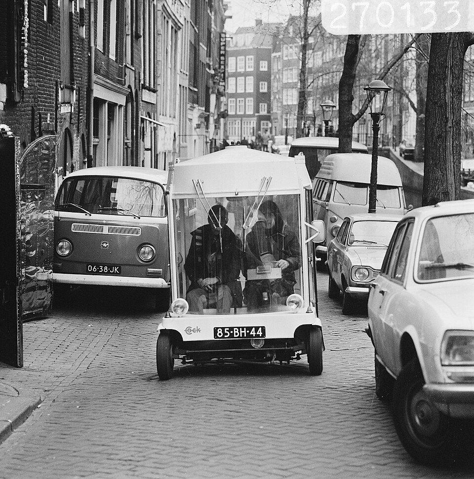 Black-and-white photograph of a glass-sided Witkar carrying two people along a narrow Amsterdam street between parked cars.