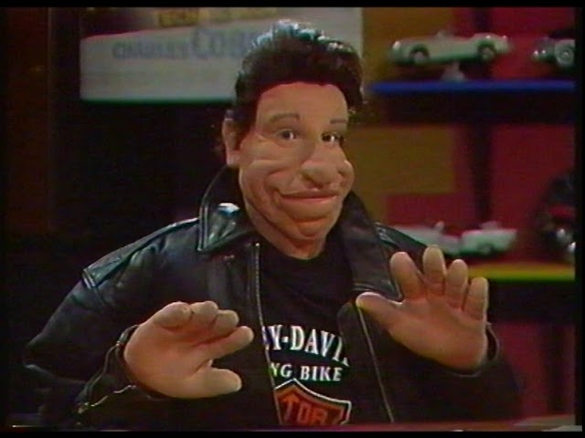 Vito, a puppet with slick dark hair and exaggerated facial features, gestures in a black leather jacket in the Game Over studio.
