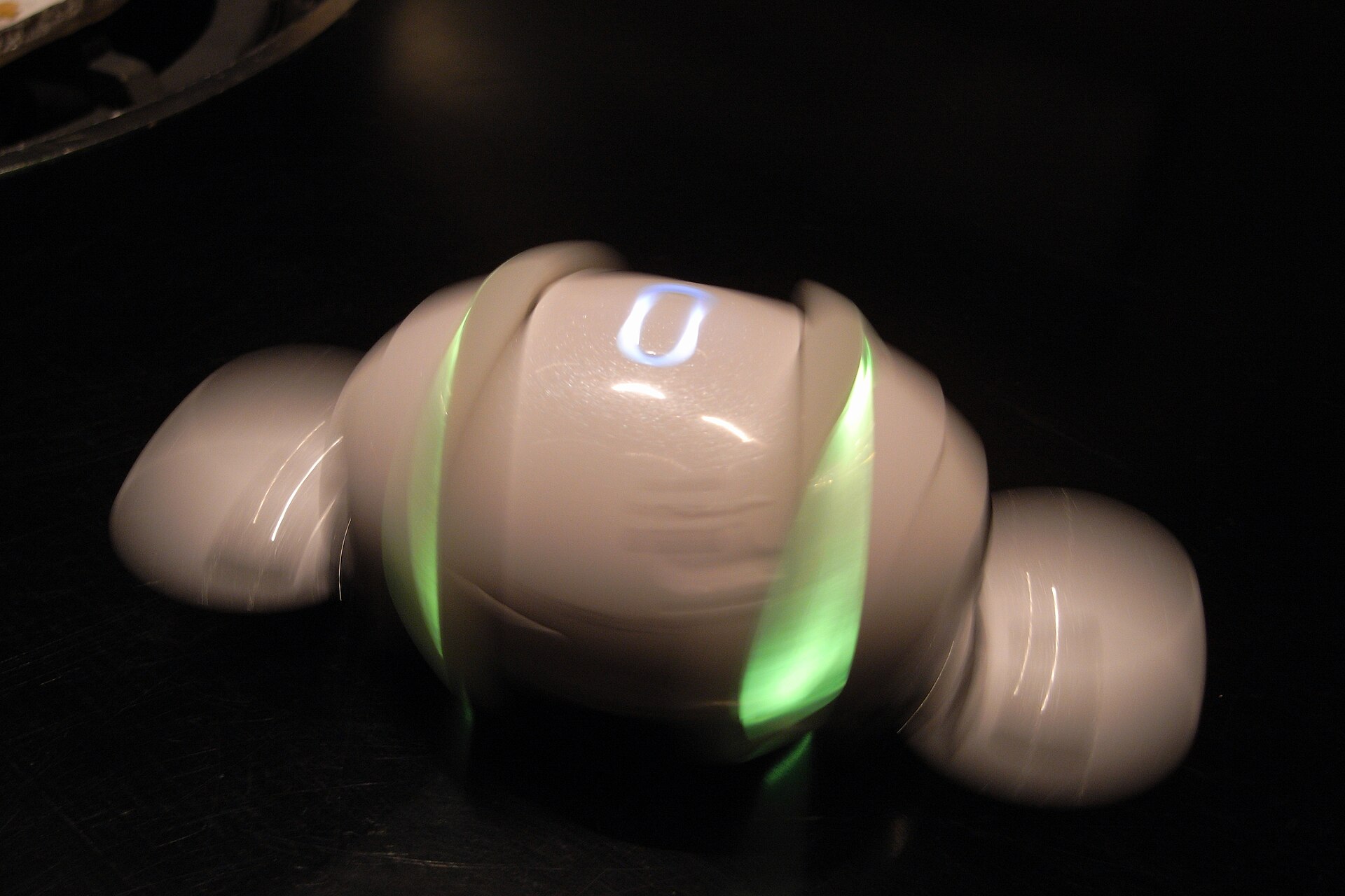 White egg-shaped Sony Rolly with green illuminated rings and open end flaps, blurred in motion against a dark surface.