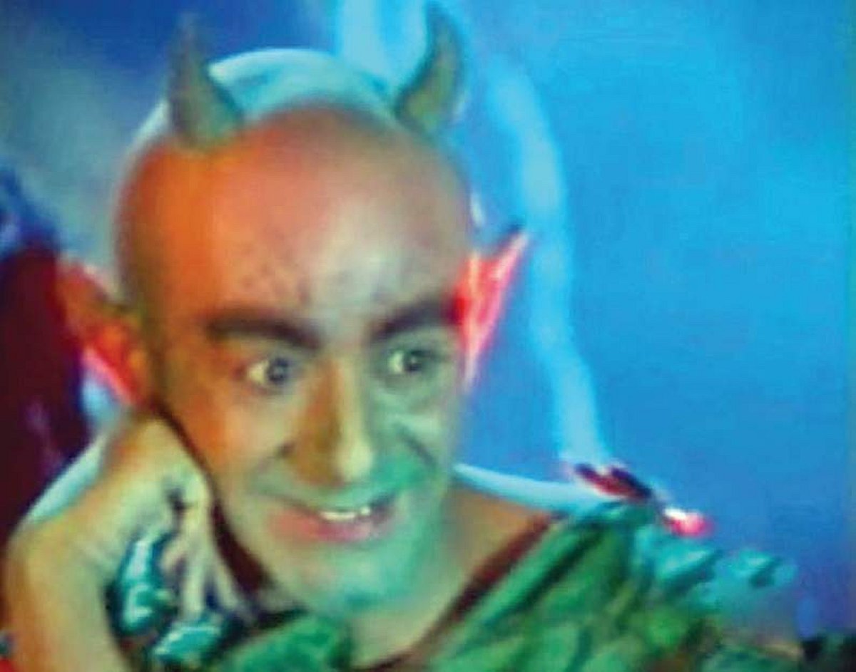 Close-up of a smiling horned mascot in a ragged green costume, with pointed ears and one hand raised beside his face against blue lighting.
