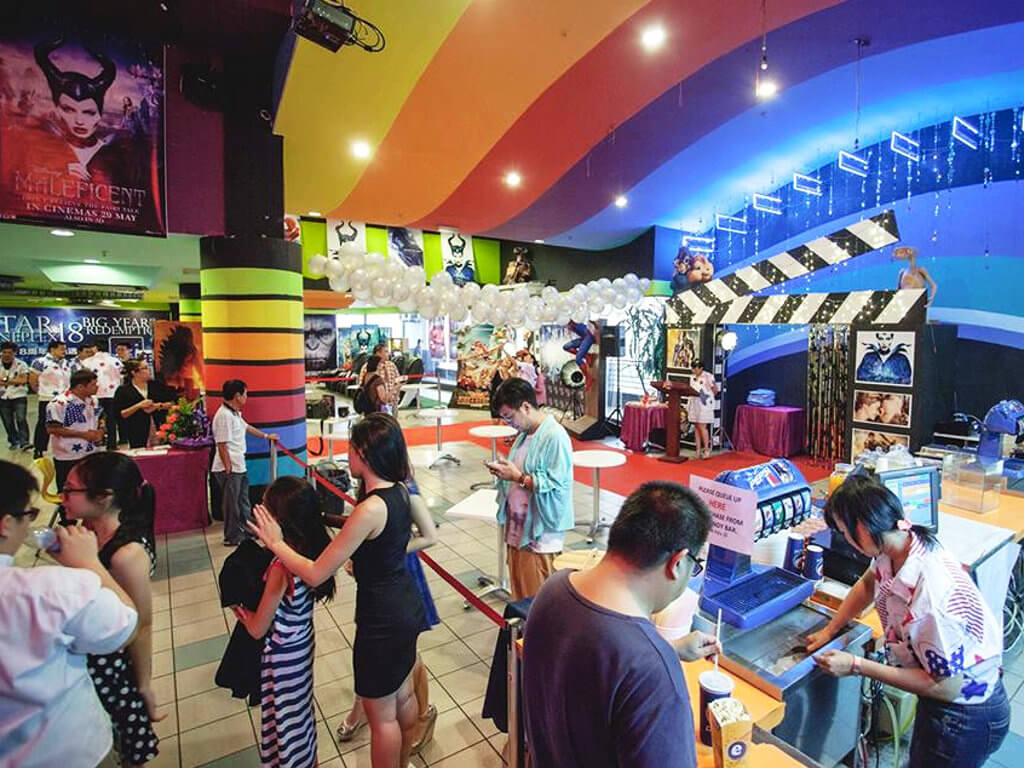 Busy cinema lobby with patrons queueing at a concession counter beneath a ceiling painted in broad rainbow-coloured bands; film posters, balloons and a large clapperboard-shaped display fill the background.