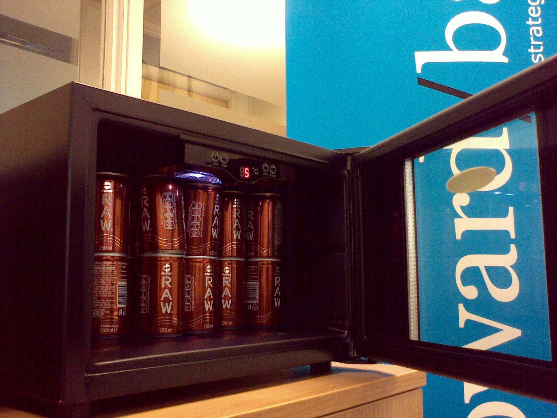 Bronze Pepsi Raw cans marked 150 ml are stacked inside a small open refrigerator beside a blue Howard Baines sign.