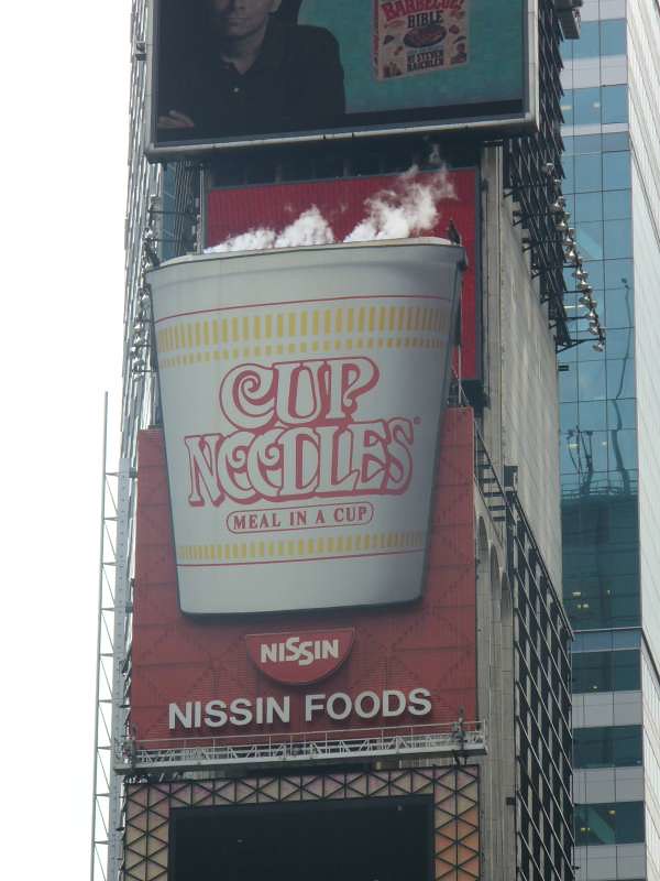 A large Cup Noodles cup projects from a red Nissin Foods billboard on a Times Square building, with white steam rising above the rim and an electronic screen overhead.
