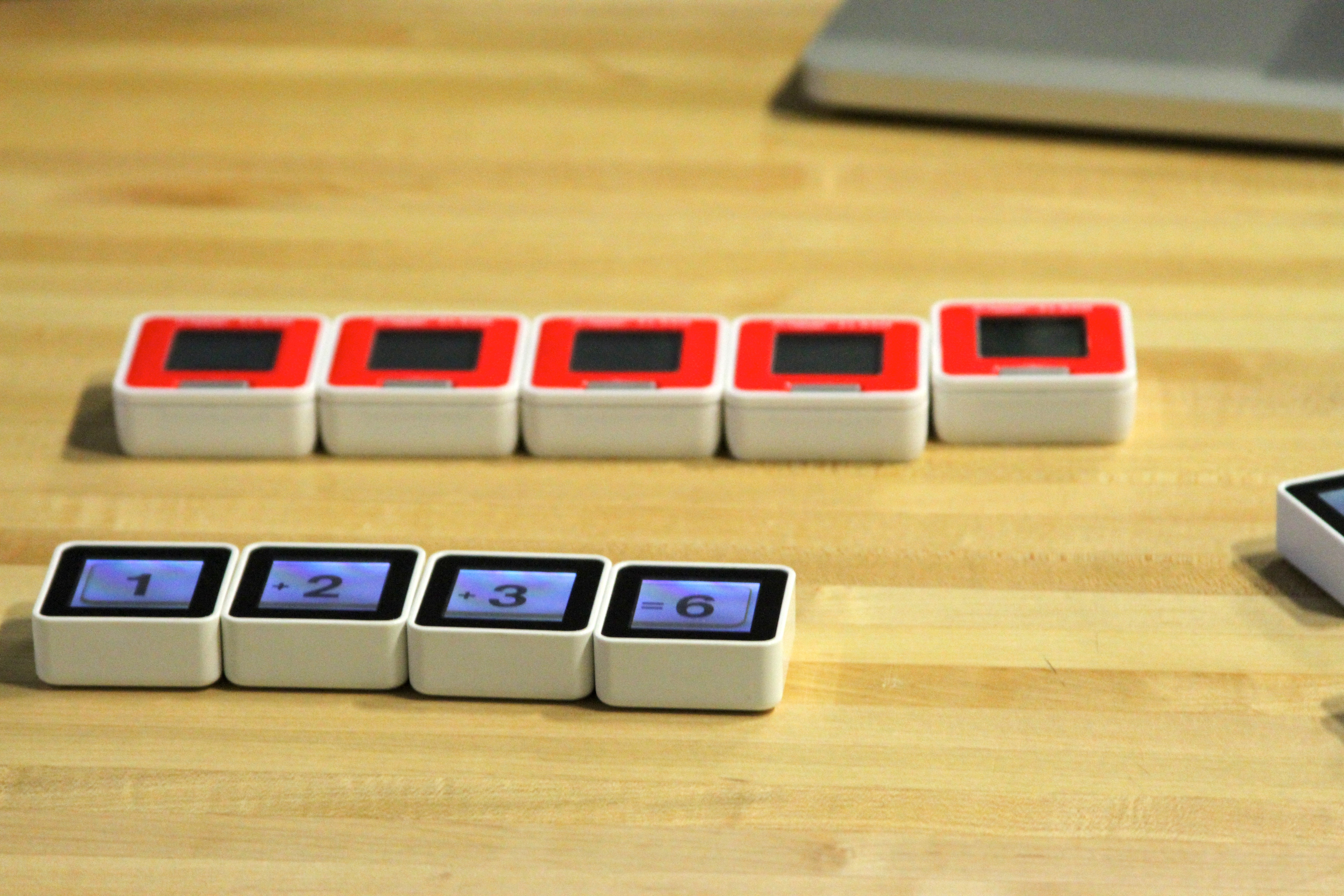 On a wooden table, four white screen blocks show1,+2,+3 and=6, with five red-topped blocks behind and part of another screen block at the right edge.