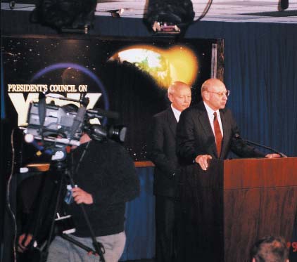 Thomas Pickering stands at a wooden podium with John Koskinen behind him, a television camera operator in the foreground and President’s Council on Y2K backdrop.