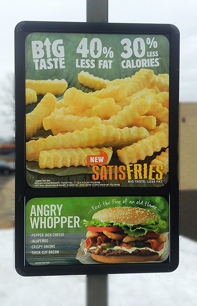 A drive-through sign shows a green Satisfries panel with crinkle-cut fries, large fat and calorie reduction claims and small comparison text. A separate Angry Whopper advertisement fills the lower panel.