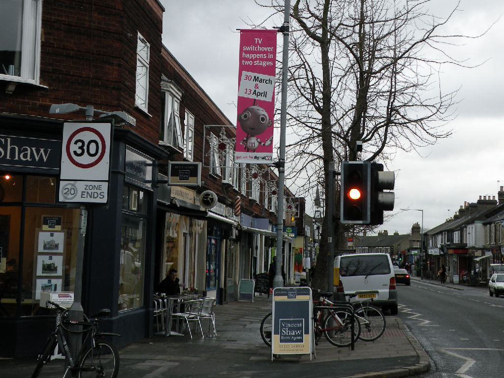 A pink street banner above Cambridge shops shows the grey Digit Al robot and TV switchover dates of 30 March and 13 April.