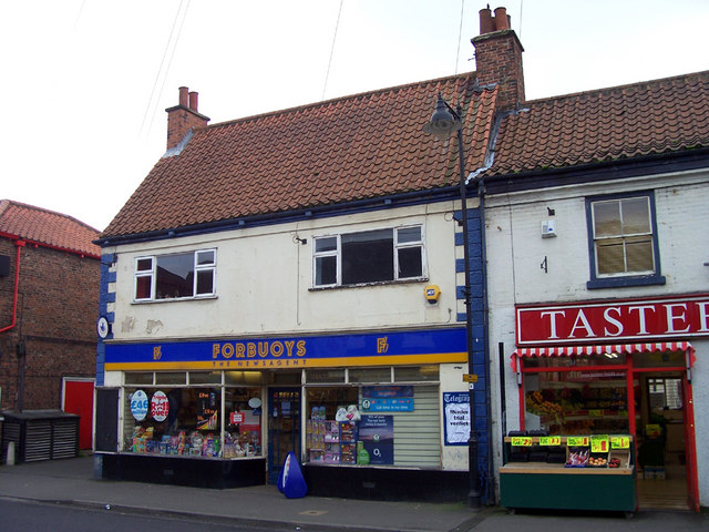 A two-storey tiled-roof building with a blue and yellow Forbuoys Newsagent sign above two shop windows, beside a red-signed neighbouring shop.