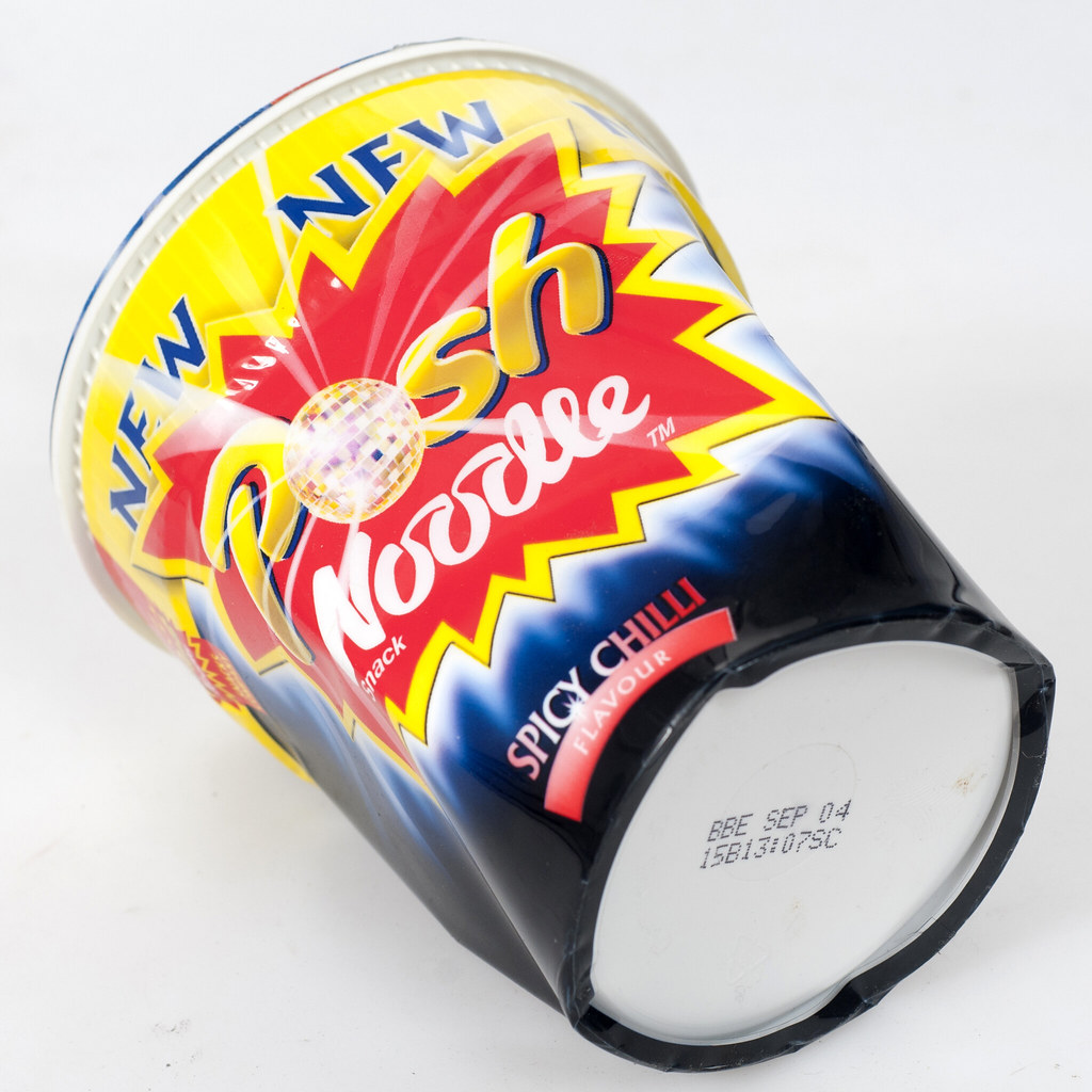 Crumpled black Posh Noodle pot on its side against white, with bright yellow and red Spicy Chilli branding and a base stamped BBE SEP 04.
