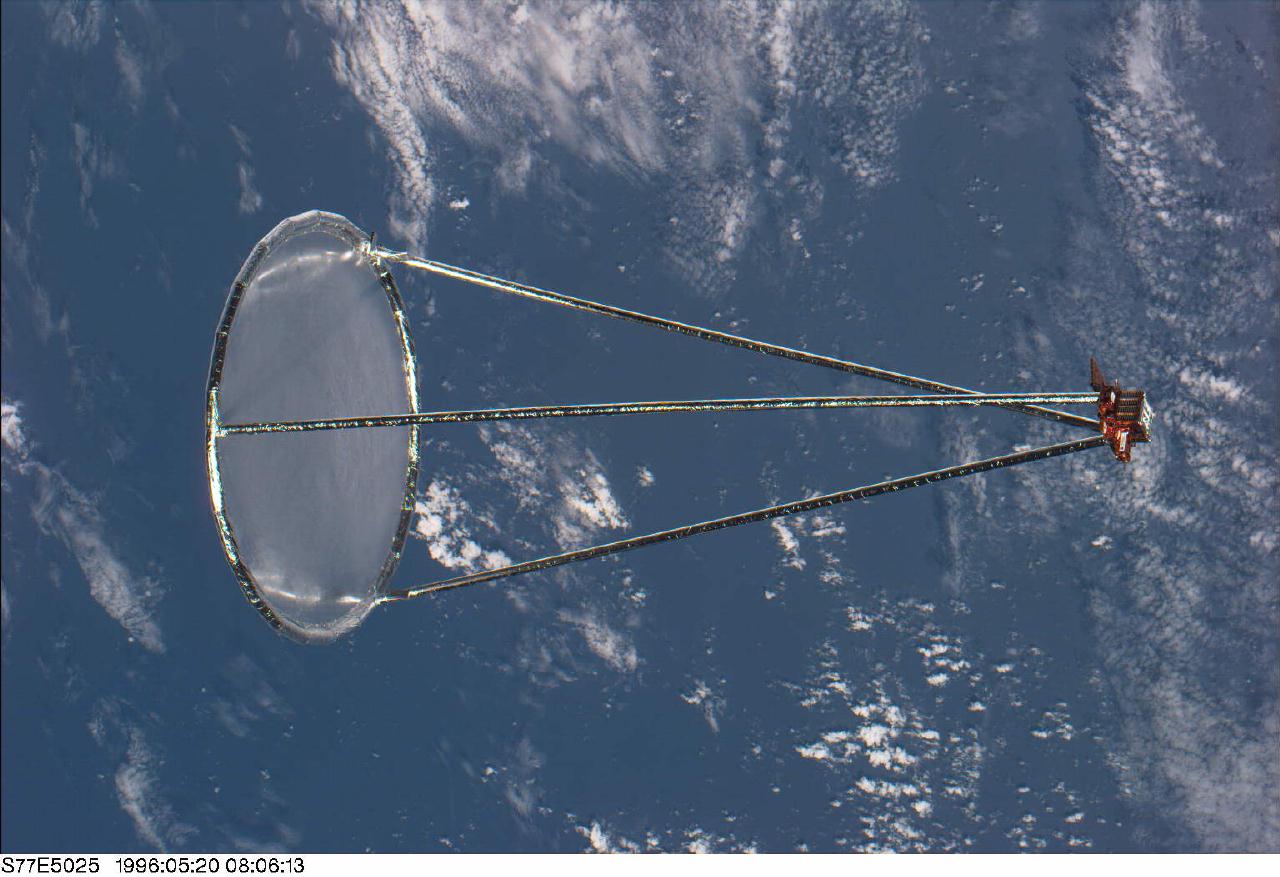 A translucent oval dish and three long slender supports stretch across a view of Earth and clouds, with a small equipment unit at the right.