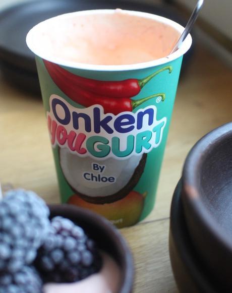 Open Onken YouGurt pot labelled “By Chloe”, decorated with fruit and chilli illustrations, with a spoon inside and a bowl of blackberries nearby.