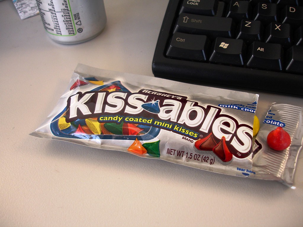 An opened silver Hershey’s Kissables packet lies on a desk beside a black keyboard, with a red sweet at the right and a drinks can behind it.