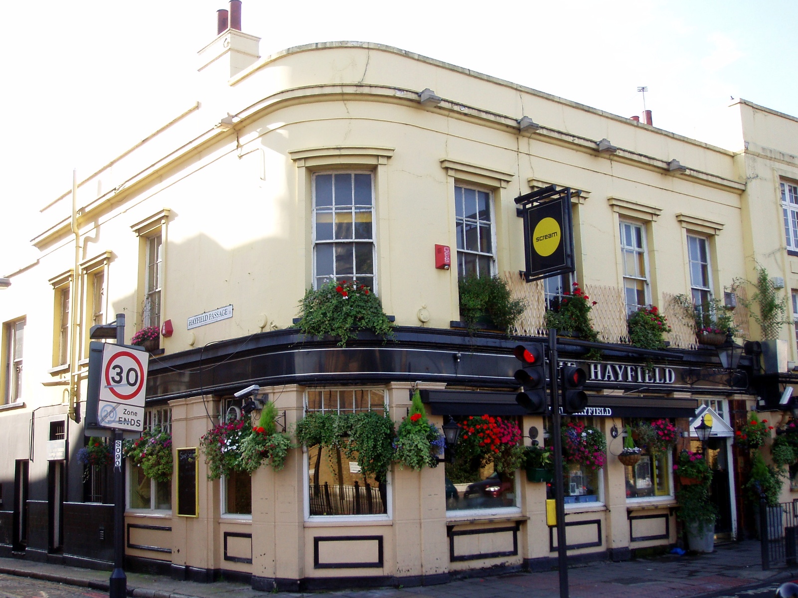 Cream-painted corner pub named Hayfield on Mile End Road, with hanging flower baskets and a black sign containing Scream in a yellow circle.