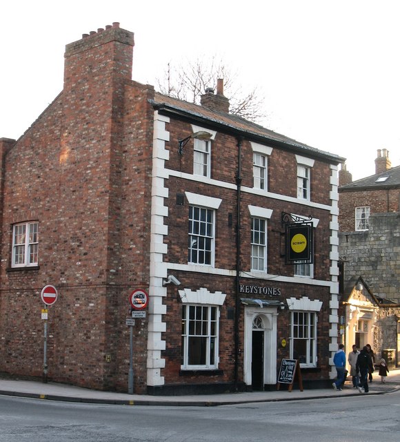 Three-storey red-brick Keystones pub with white stone window surrounds, a black hanging sign with a yellow Scream circle, and pedestrians beside the entrance.