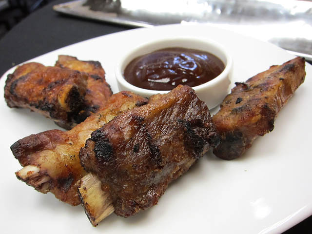 Several browned bone-in ribs sit on a white plate around a small bowl of dark barbecue sauce.