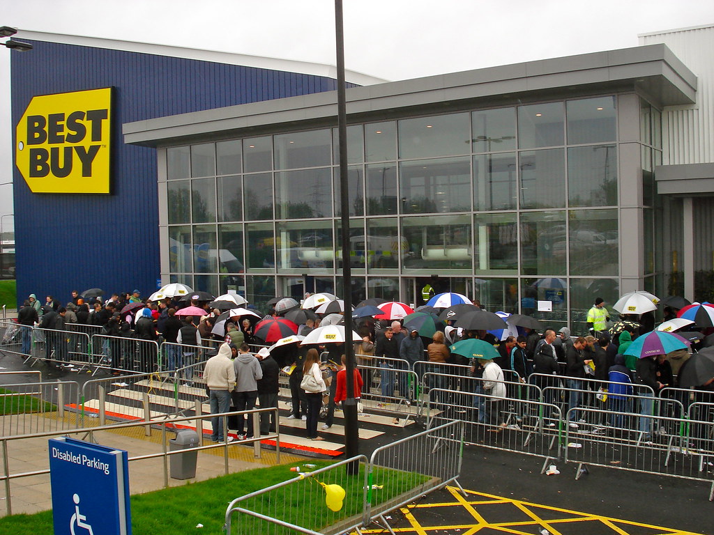 People shelter under umbrellas in a barrier-lined queue outside a glass-fronted shop with blue cladding and a large yellow Best Buy sign. A car park and accessible-parking sign fill the foreground.