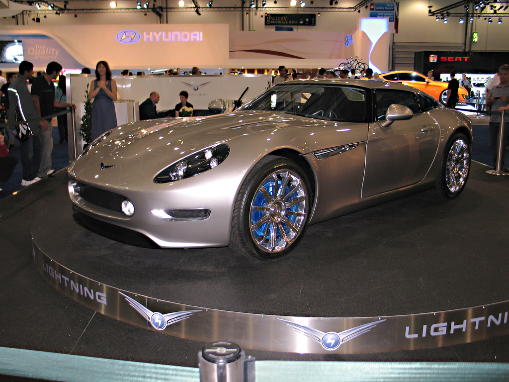 Silver two-door Lightning GT on a round black display platform, photographed from the front left amid exhibition visitors and stands.