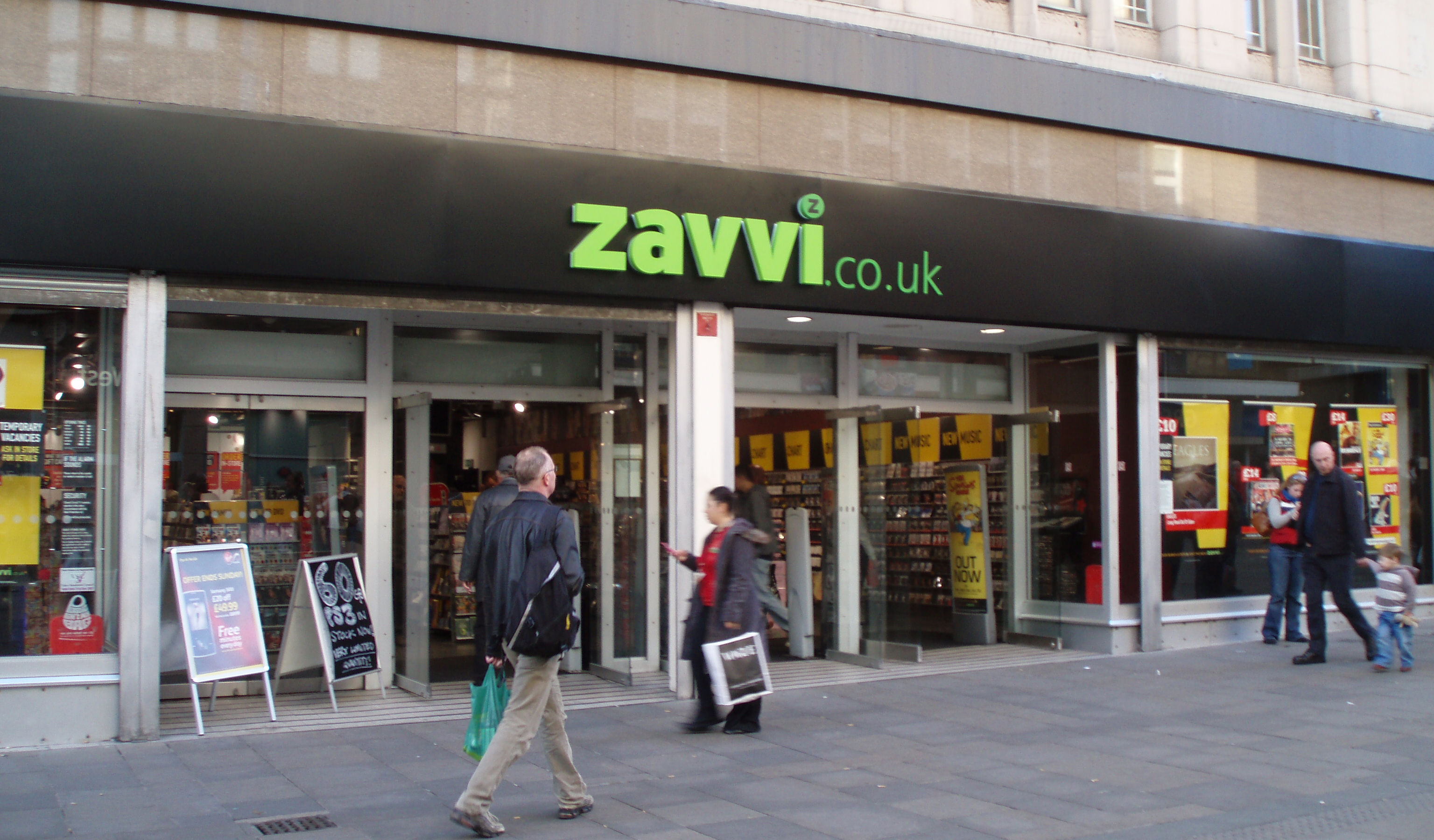 Bright green zavvi.co.uk lettering spans a black fascia above an open music shop, with racks and yellow posters inside and pedestrians passing outside.