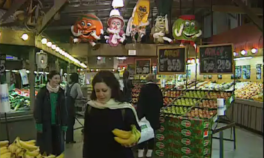 Shoppers stand among produce displays beneath five large cartoon food and family figures mounted above a supermarket aisle.
