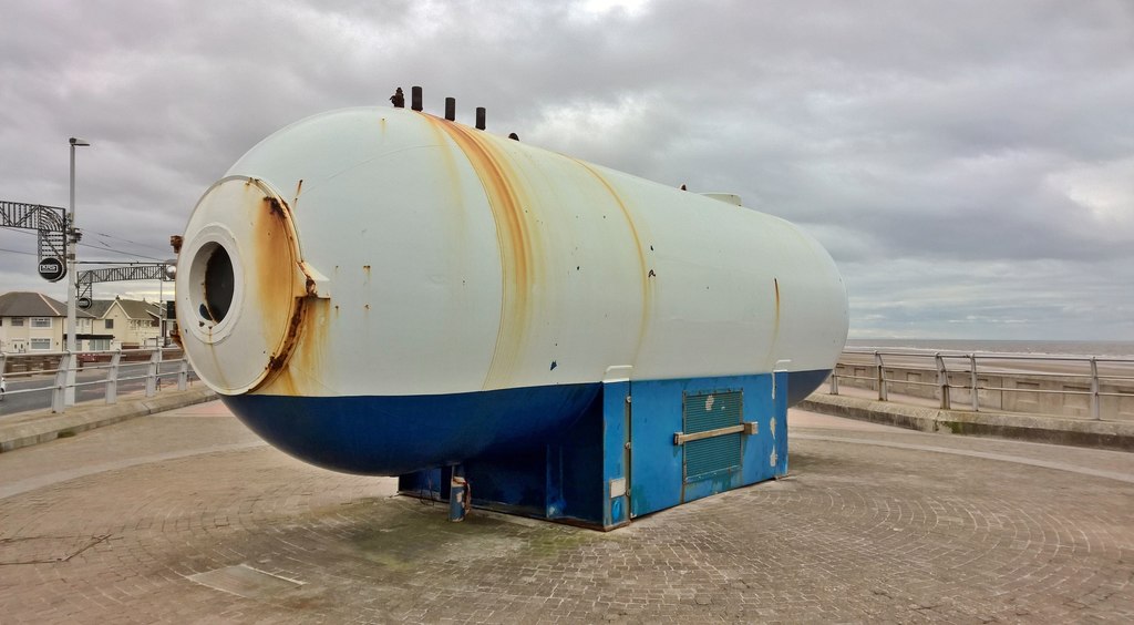 A pale cylindrical metal chamber with a blue lower band, round porthole and rust streaks stands on the paved promenade beside the sea.