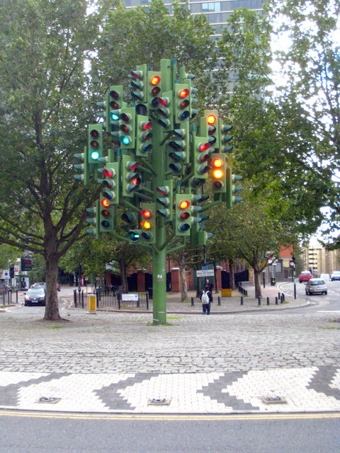 Branchinggreenmetalpolescarrymanylitred,amberandgreensignals;livingtrees,buildings,traffic,pedestrianandroundaboutpavingsurroundthetrunk.