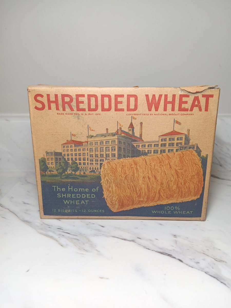Front of a worn beige Shredded Wheat cereal box on a white marble surface. Large red lettering, an illustrated factory, and a large brown shredded-wheat biscuit appear on the carton; smaller text includes “The Home of Shredded Wheat,” “12 biscuits—12 ounces,” and “100% whole wheat.”