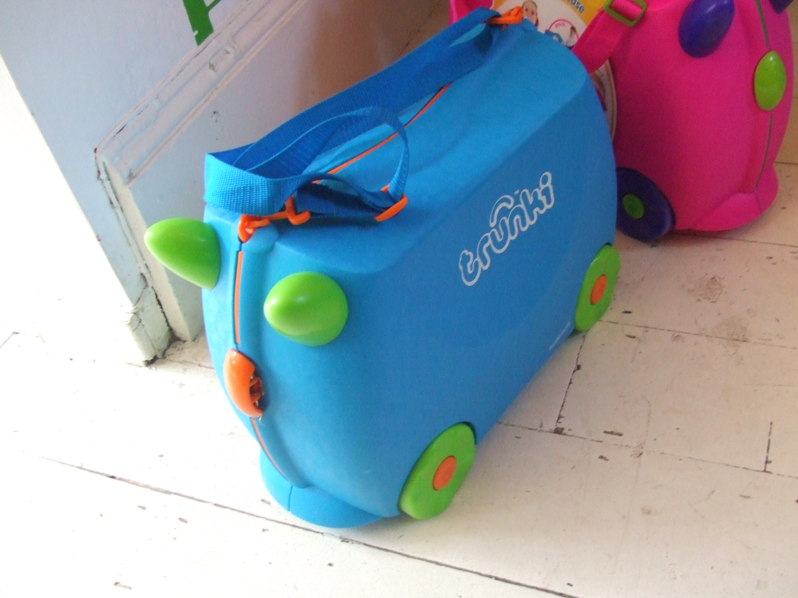 A blue Trunki suitcase with green wheels and horn-shaped handles, beside a pink suitcase.