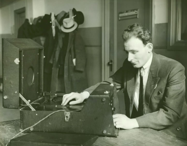 A worker in a suit tests an open portable talking-book record player at a table.