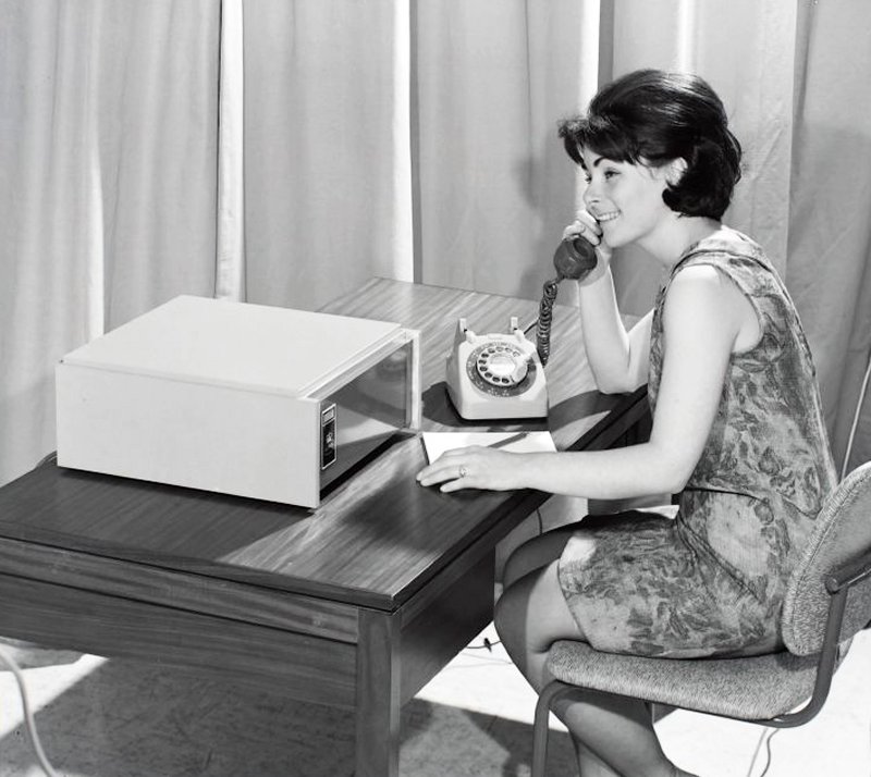 Woman at an office desk holds a telephone receiver beside the grey cabinet of a Datel modem.