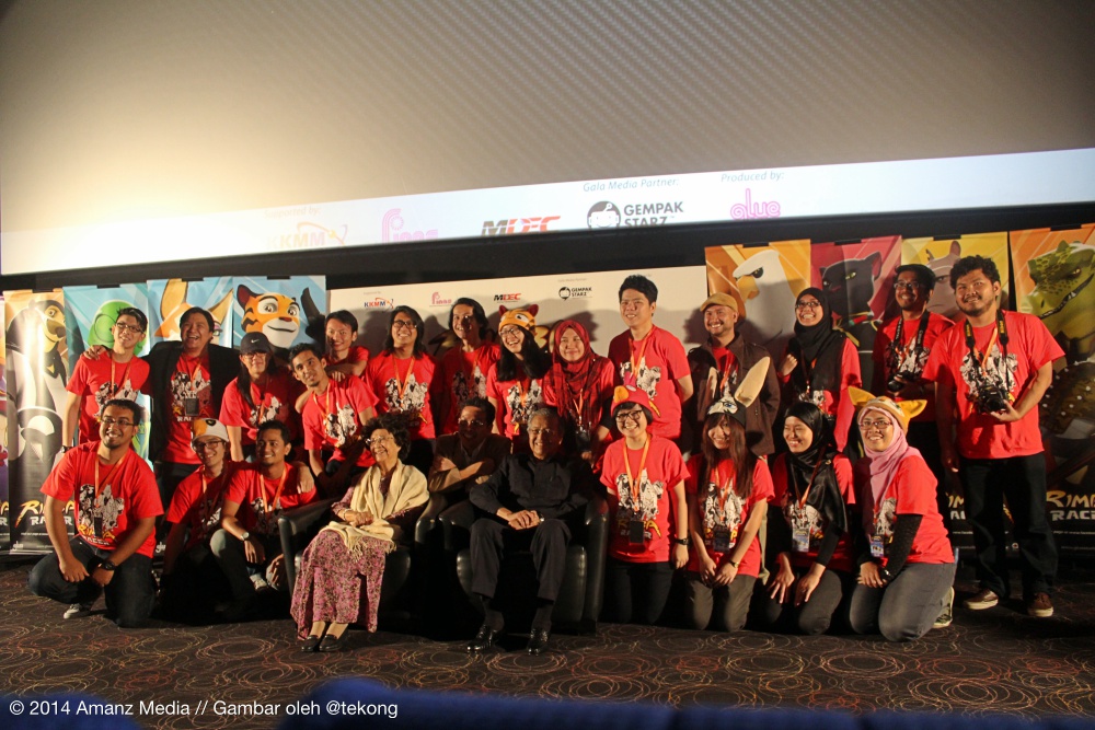 A large group poses in rows at a launch venue, many wearing red Rimba Racer shirts, with animated animal character banners and production-partner logos behind them; an Amanz Media/@tekong watermark appears at bottom left.