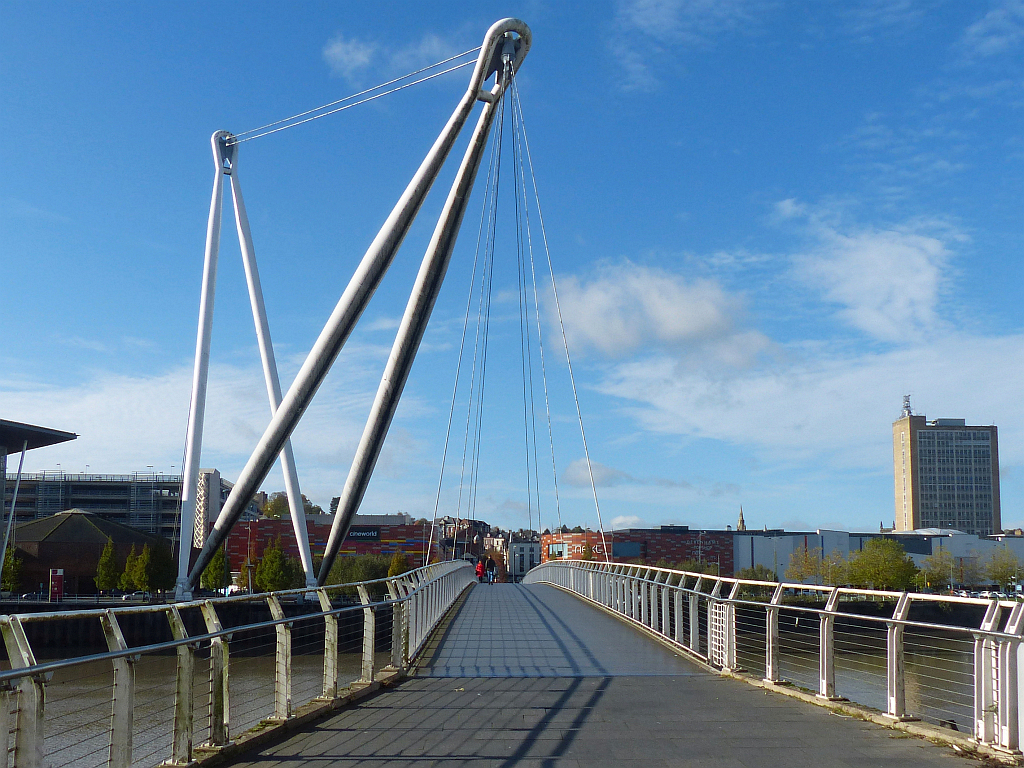 A paved footbridge deck leads toward city buildings beneath tall angled white masts and suspended cables against a blue sky.