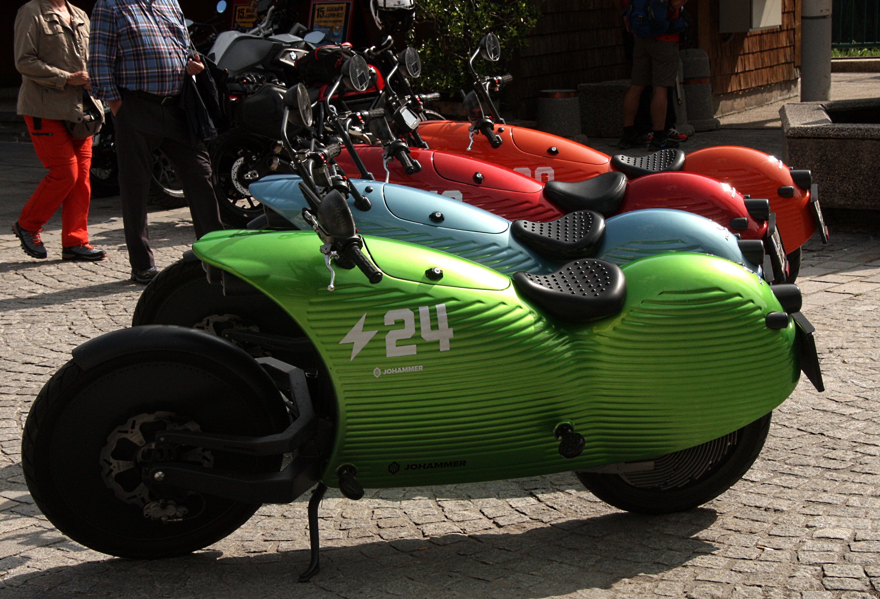 Four ribbed electric motorcycles in green, blue, red and orange stand diagonally on cobblestones, with high mirrors, perforated black seats and people behind them.