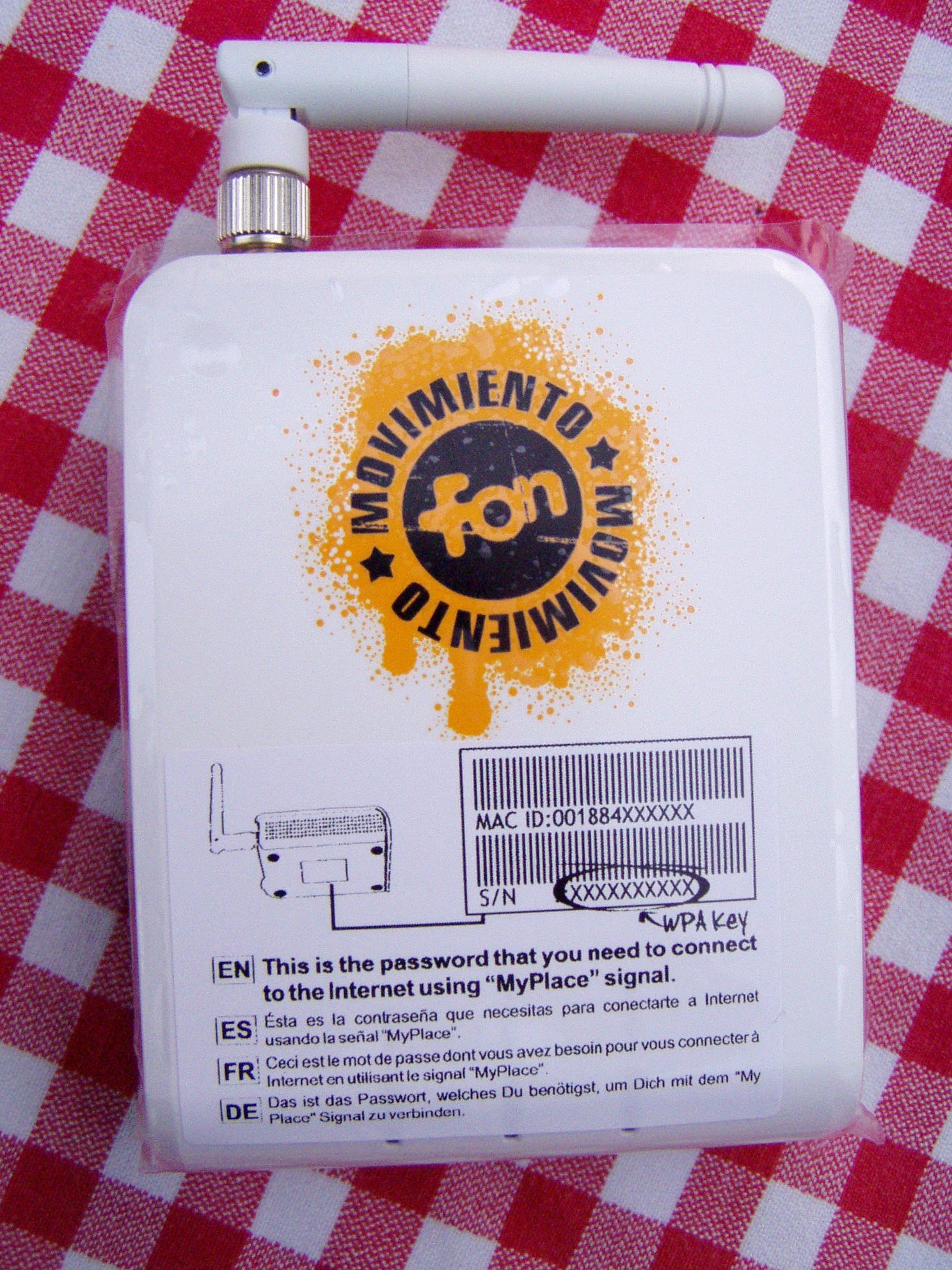 A white La Fonera router with a folded antenna rests on red-and-white checked cloth, bearing an orange FON Movimiento logo and a multilingual protective label explaining the MyPlace password.