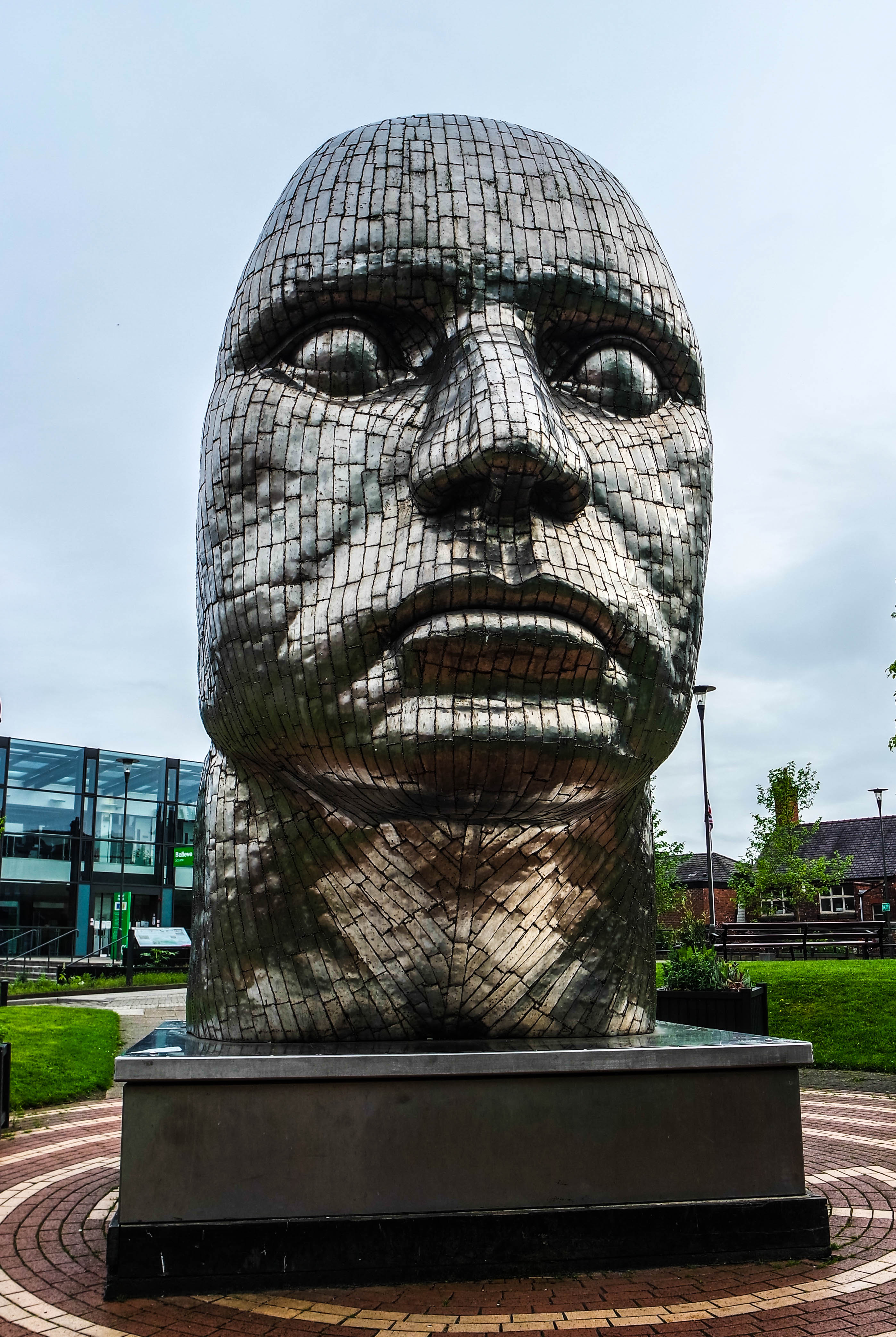 A tall metallic face assembled from small welded plates stands on a rectangular base beside lawns and the glass-fronted Wigan Life Centre.