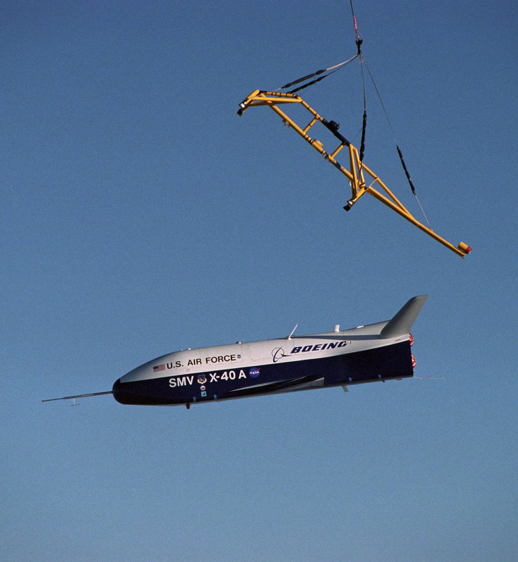 A small white and black X-40A aircraft marked U.S. Air Force, Boeing and NASA flies below a suspended yellow lifting frame against a blue sky.